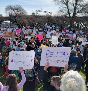 Protesters at a rally. Two kids hold signs in the foreground. One says, Strong Girls Strong Future and the other says BE FAIR TO WOMEN
